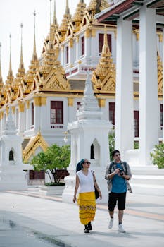 Two tourists walk by a golden Thai temple with intricate architecture in daylight.