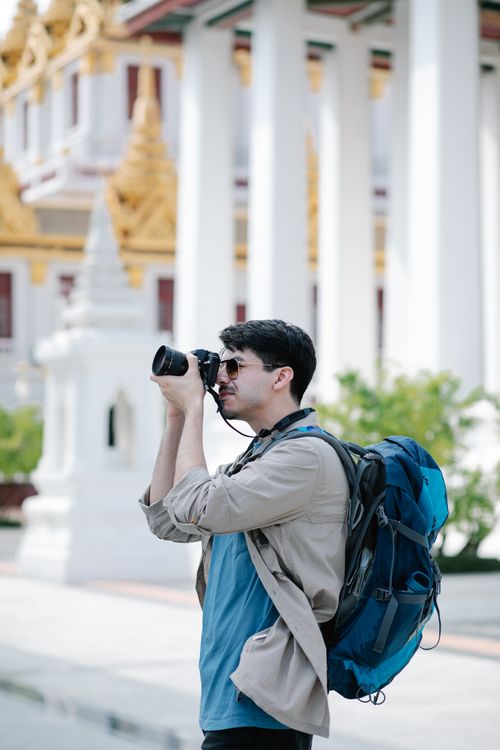 Free Man taking a photo at a temple outdoors, carrying a blue backpack and wearing sunglasses. Stock Photo