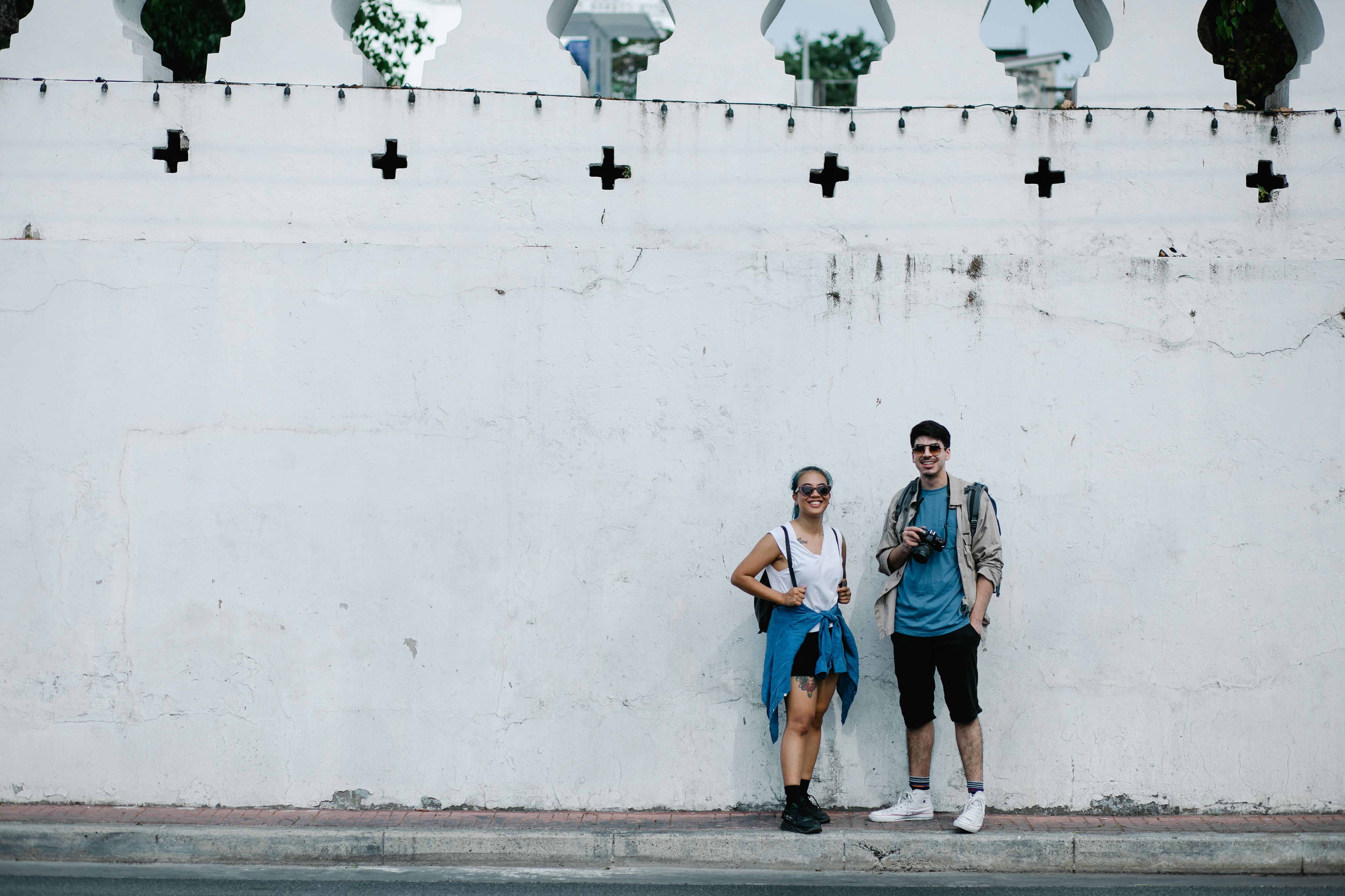 A happy couple in casual wear posing against a historic white wall during their travels.