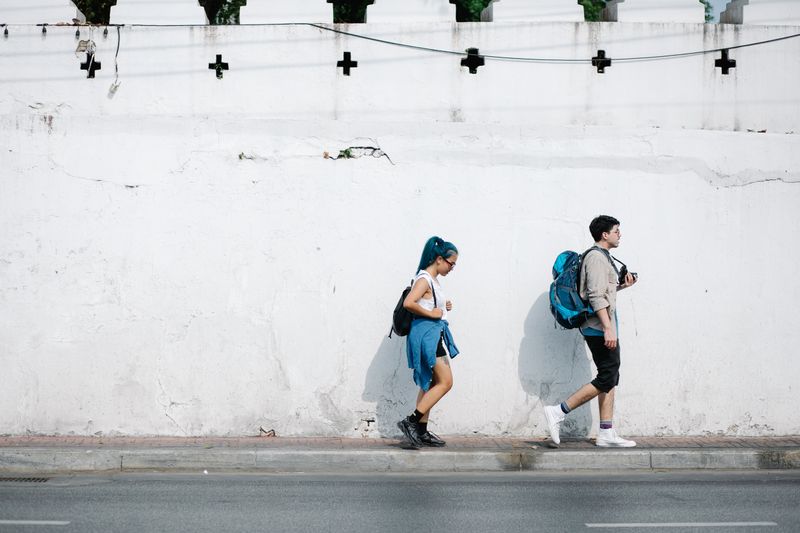 Young adults with backpacks walking on sidewalk against a white wall in an urban setting.