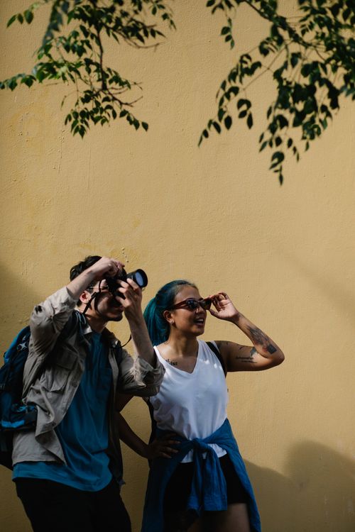 Free A man with a camera and a woman posing against a vibrant yellow wall, enjoying a lively photoshoot. Stock Photo