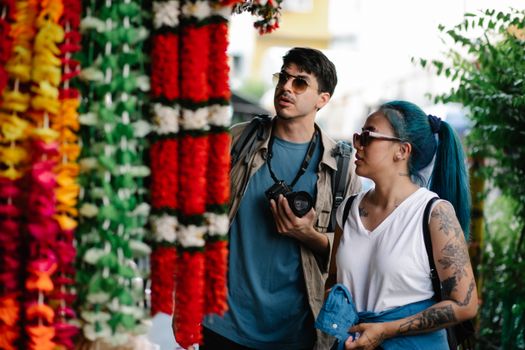 A couple explores a vibrant outdoor market, admiring colorful flower garlands.
