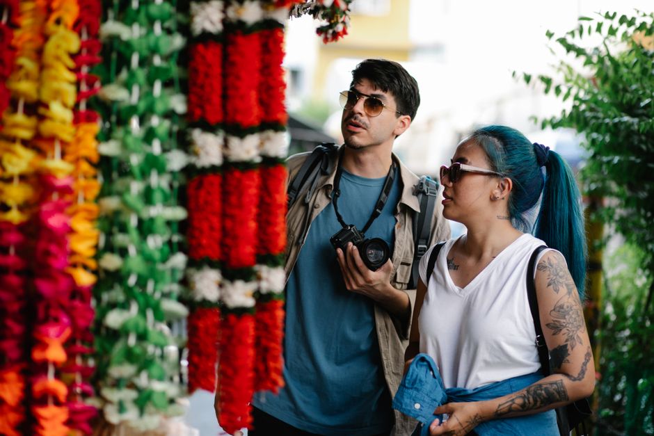 A couple explores a vibrant outdoor market, admiring colorful flower garlands.