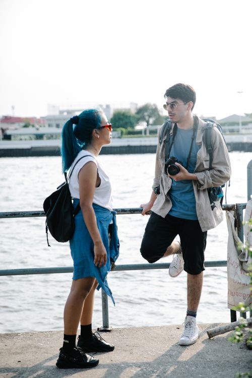 Free Two young travelers with backpacks enjoying a sunny outdoor conversation by a waterfront. Stock Photo