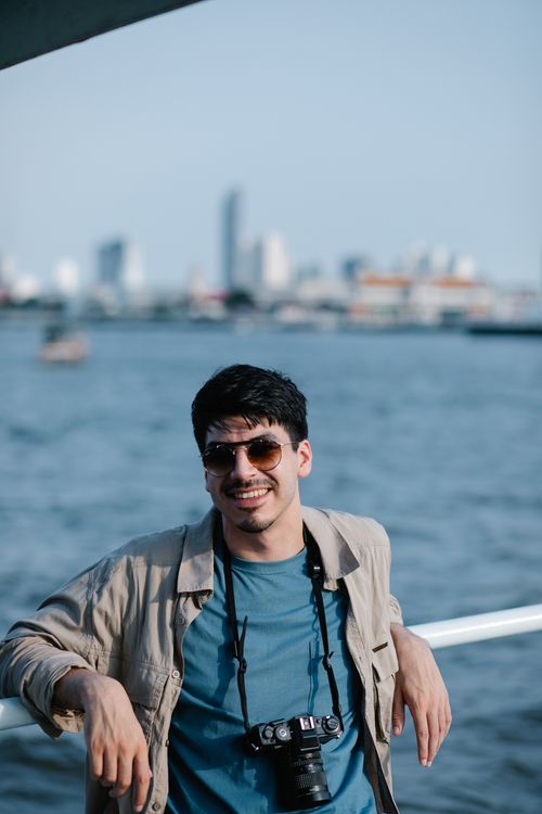 Free A cheerful man with sunglasses and a camera enjoys a sunny day by the water. Stock Photo