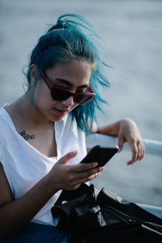 A stylish young woman with blue hair and sunglasses using her smartphone outdoors by the water.