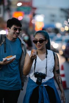 Interracial couple enjoys a street walk with food and cameras during daylight.