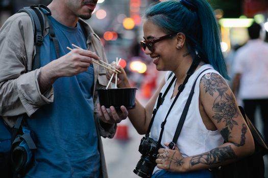 Two friends sharing a bowl of noodles with chopsticks, captured on a bustling city street.