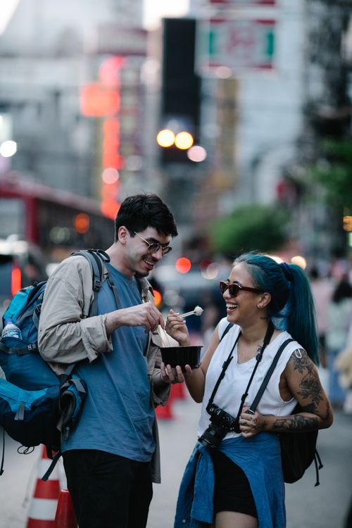 Free Young couple laughing and enjoying street food in a vibrant urban setting. Stock Photo