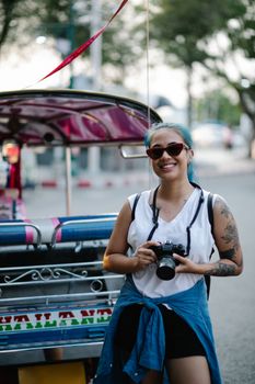 A cheerful woman holding a camera stands by a colorful tuk-tuk on a sunny day.