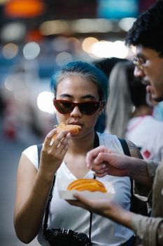 Young woman with blue hair and sunglasses eating street food in a vibrant night market setting.