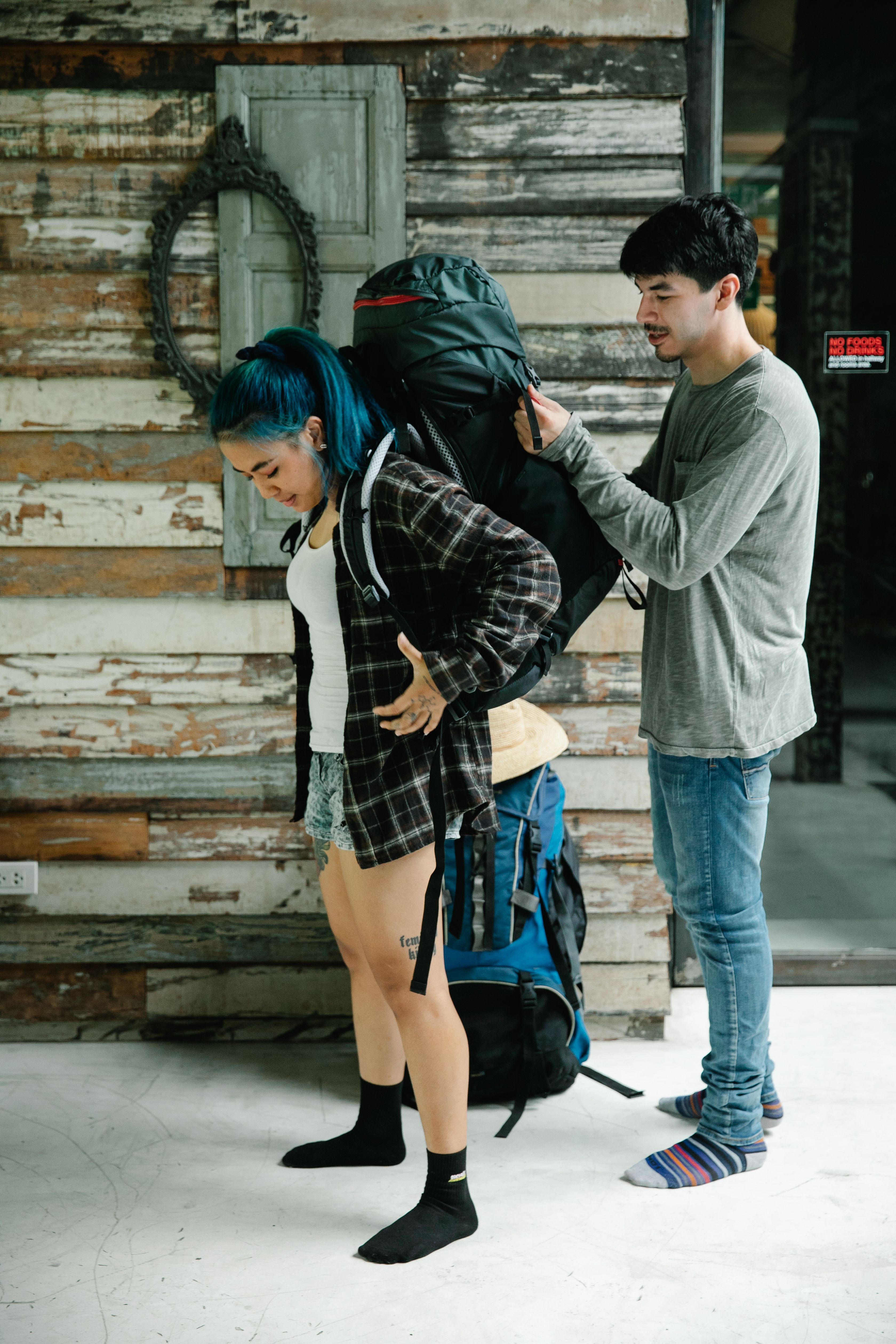 Two young adults with backpacks preparing for travel indoors, adjusting their gear.