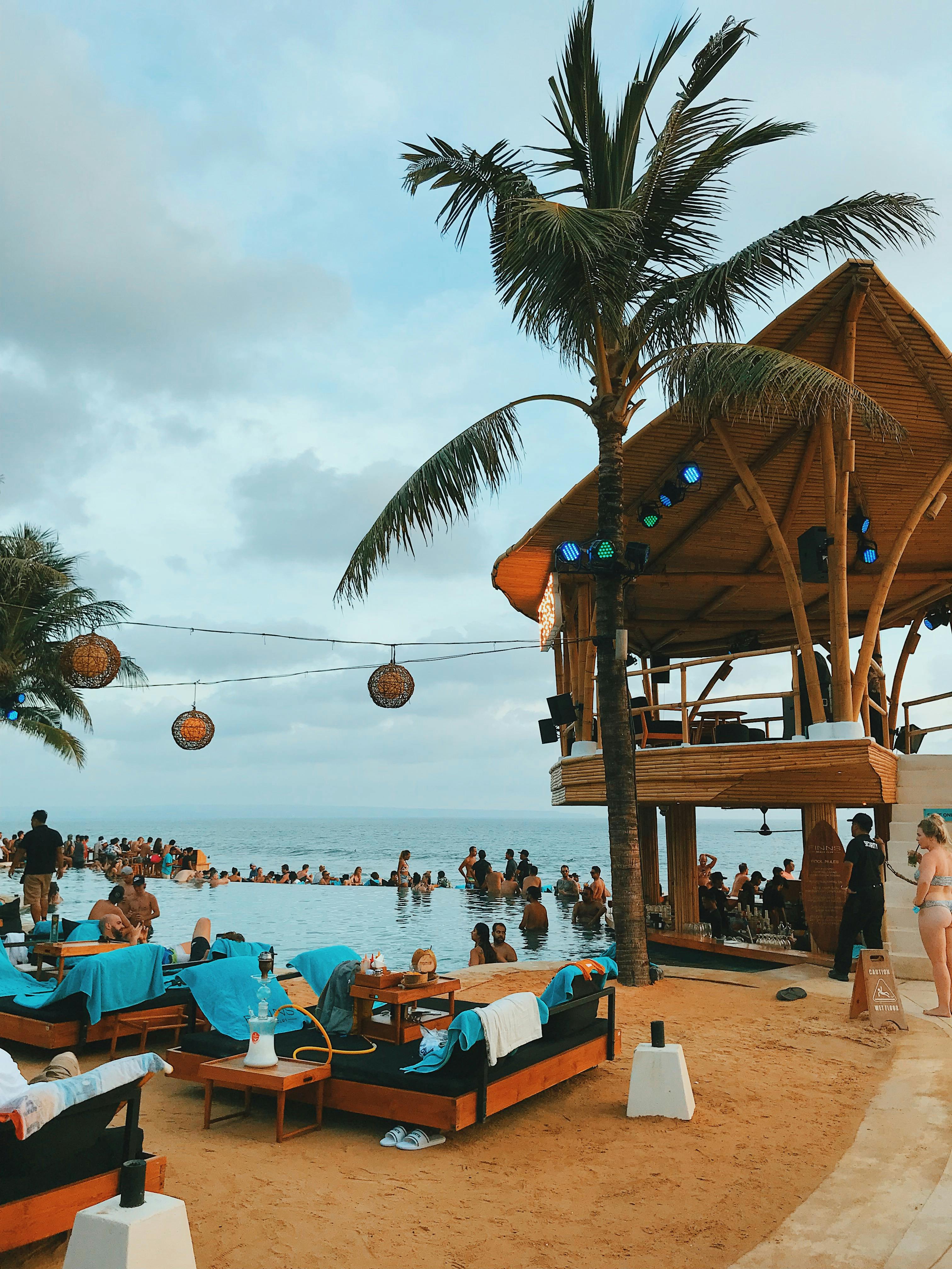 Crowd gathered at a Seminyak beach club at sunset with a DJ booth and infinity pool overlooking the Indian Ocean in Bali