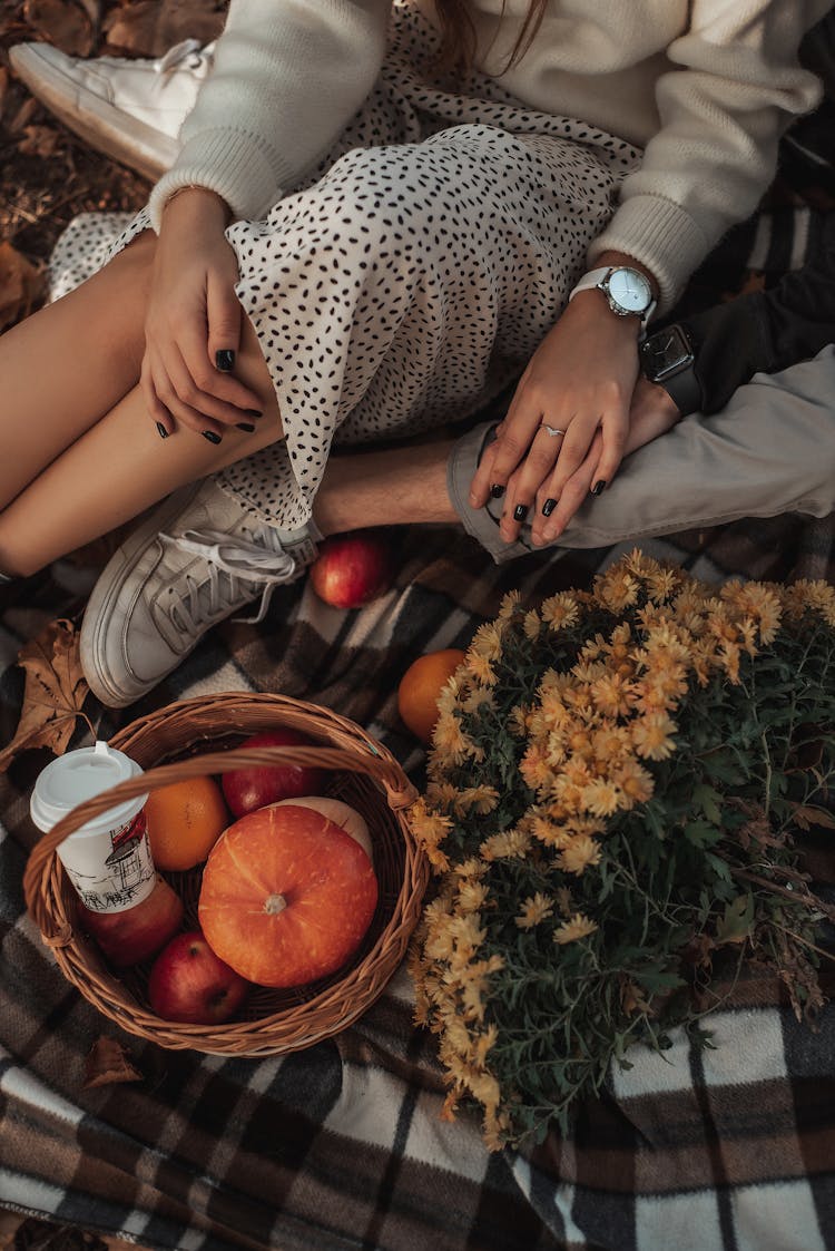 A Couple Sitting On The Picnic Blanket Beside The Bssket Of Fruits And Flowers