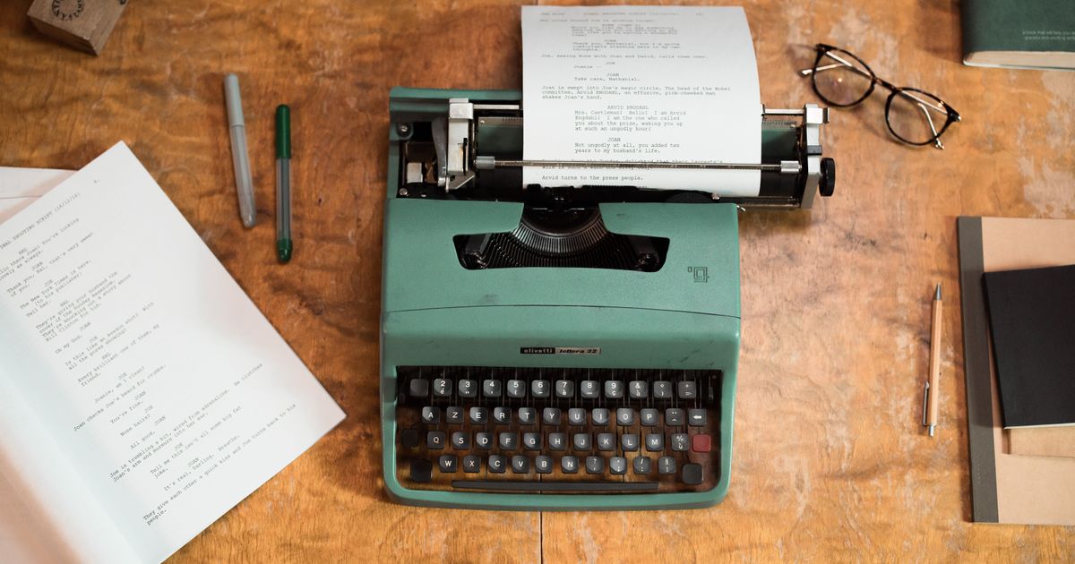 A vintage green typewriter with scripts surrounded by glasses and pens on a wooden table.