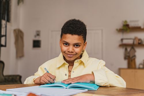 Free A young boy with curly hair smiles while studying with a notebook inside a cozy room. Stock Photo