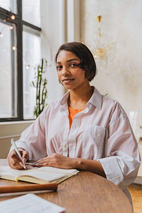 Free A young woman sitting at a table by the window, writing in a notebook. Bright and casual atmosphere. Stock Photo