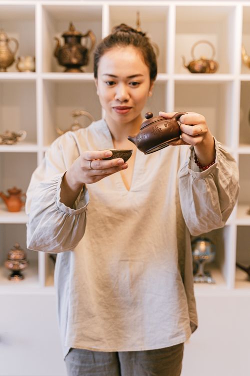 Free Asian woman pouring tea in a serene indoor setting with teapots in the background. Stock Photo
