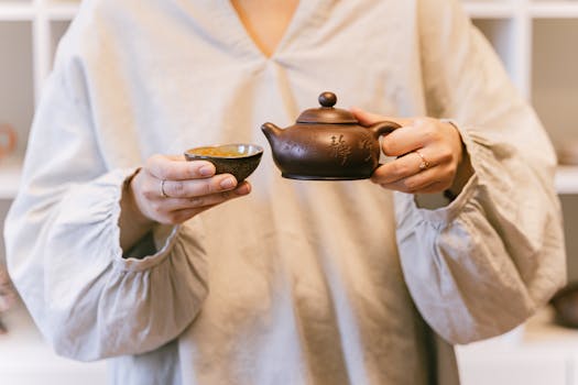 A person in white sleeves pouring tea from an antique ceramic teapot into a teacup.