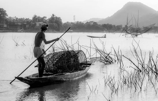 Shirtless fisherman in a canoe casting nets on a calm river in grayscale.