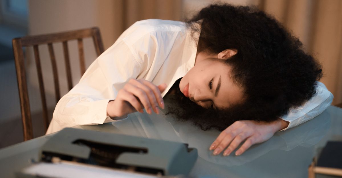 A woman with afro hair in a white shirt rests her head on a table next to a vintage typewriter, appearing fatigued.