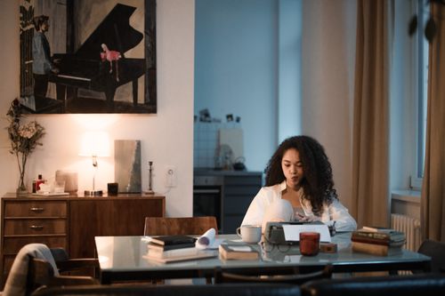 Free A woman sits at a table reading under warm indoor lighting, surrounded by books and a warm beverage. Stock Photo
