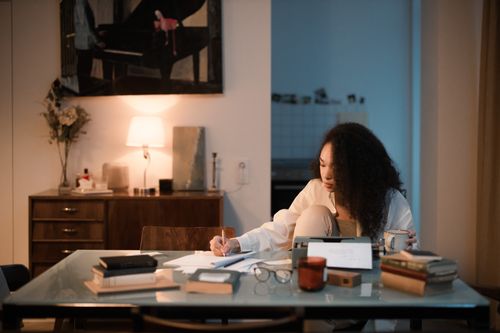 Free A woman sits indoors writing on a notepad, surrounded by books and coffee, creating a cozy workspace. Stock Photo