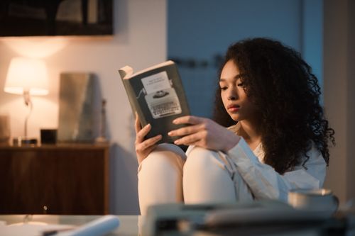 Free A young woman with curly hair enjoys reading a book in a cozy, softly lit room. Stock Photo