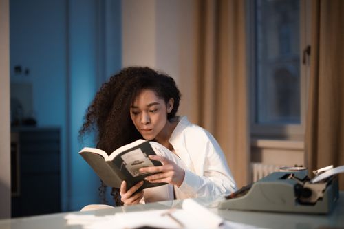 Free Young woman with curly hair engrossed in a book beside a typewriter at home. Stock Photo