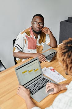 Two men engaged in a meeting with laptops and documents on a wooden desk.