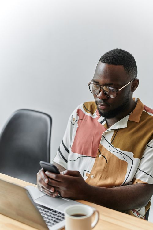 Free African American man in eyeglasses using smartphone in office setting, wearing casual shirt, with laptop and coffee. Stock Photo