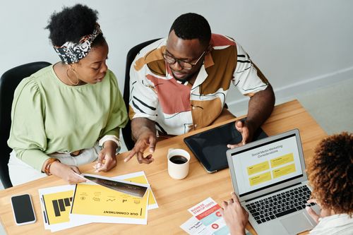 Free Three colleagues collaborate over marketing documents and a laptop in a modern office setting. Stock Photo