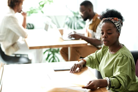 A focused woman working in a modern office environment with colleagues in the background.