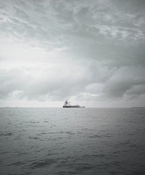 A lone cargo ship sails on the vast ocean under a dramatic cloudy sky in grayscale.