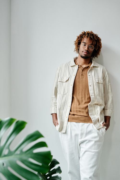 Free Portrait of a stylish African-American man with curly hair and glasses leaning against a white wall indoors. Stock Photo