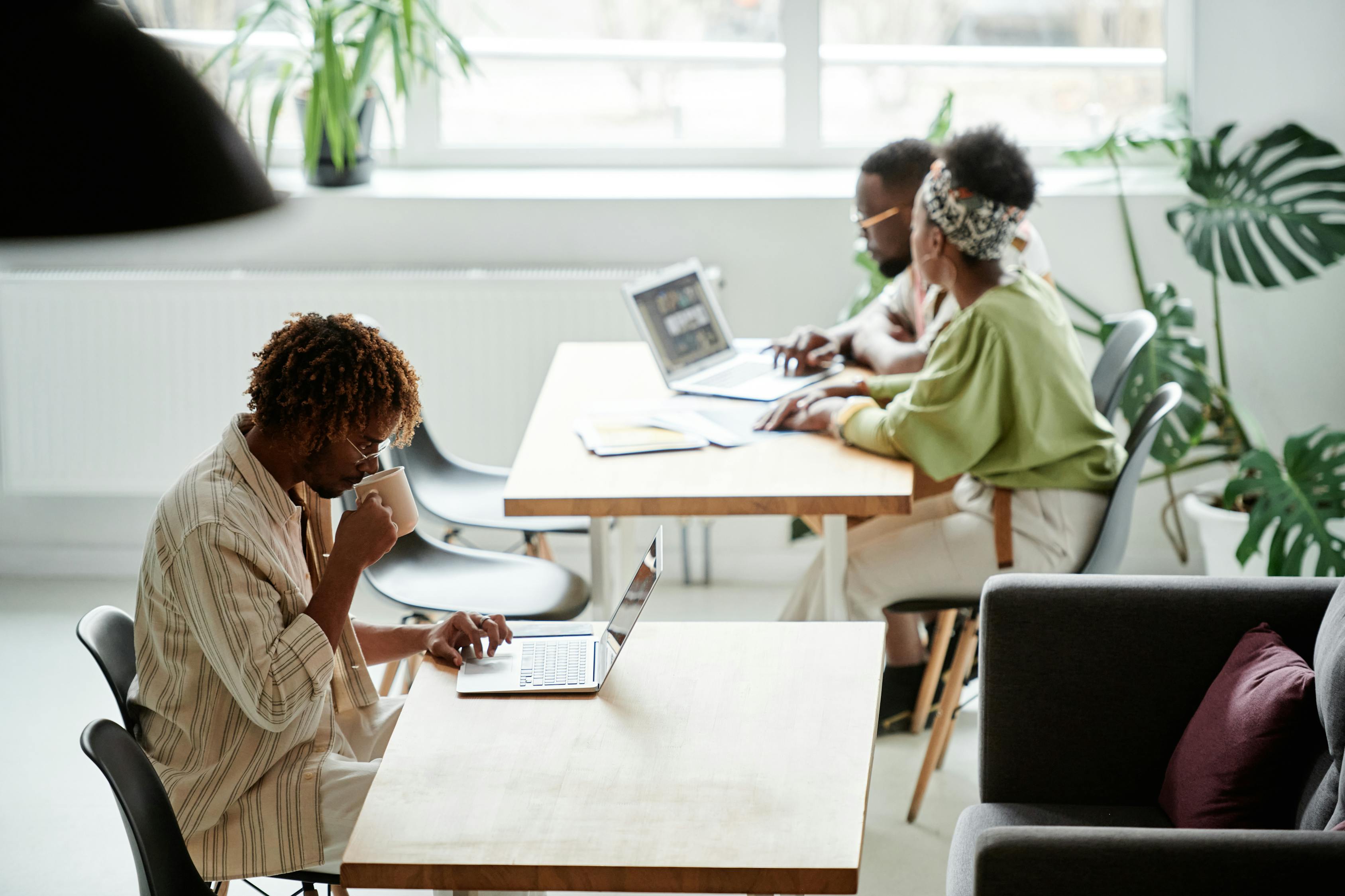 man drinking from mug at a table with laptop and two people sat at another table working on laptop