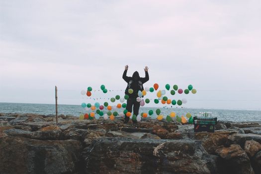 Unrecognizable male seller with raised arms in hood standing with row of balloons for playing darts on stony waterfront