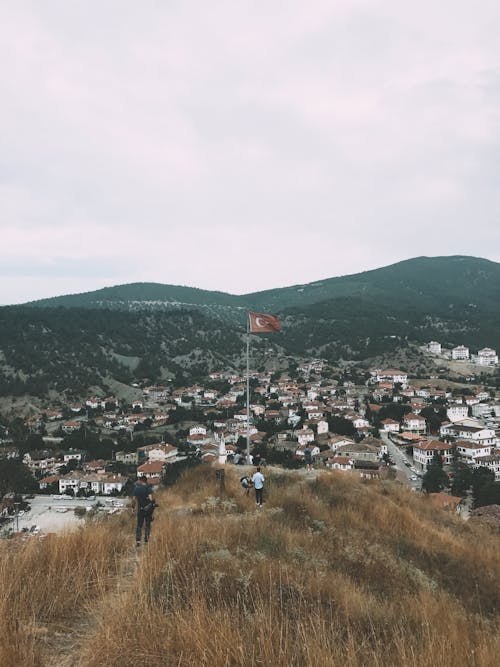 Distant people standing on grassy hilltop with waving Turkish flag hanging on flagpole above town