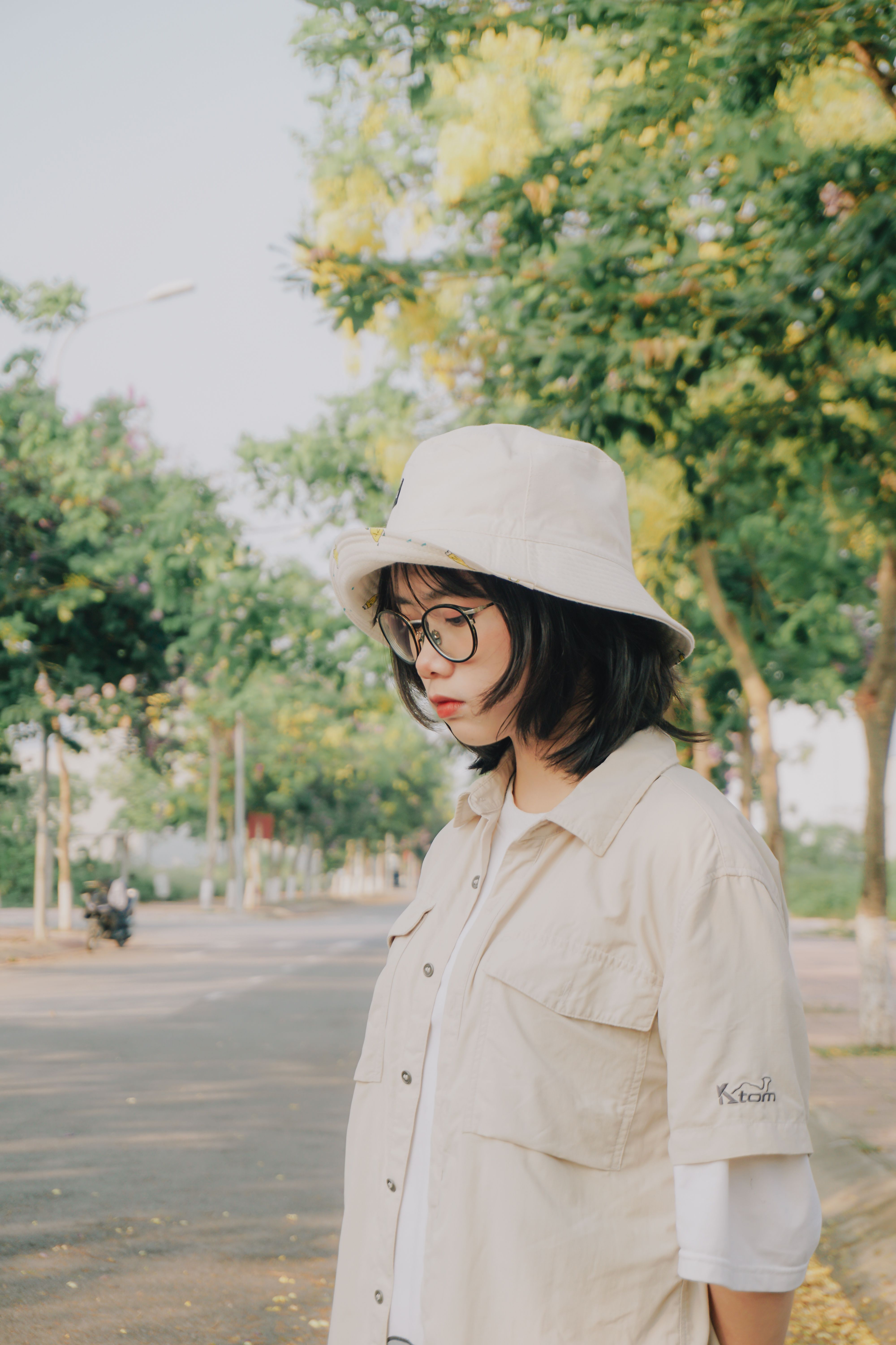 A young woman wearing a hat and eyeglasses walks on a tree-lined street in Vietnam.