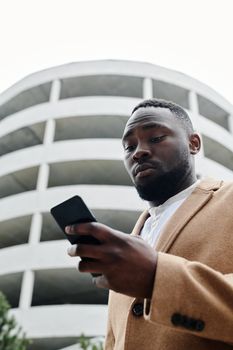 A young businessman looks at his smartphone in front of a circular building, dressed in a stylish brown coat.