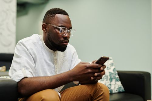 Free A man wearing glasses and brown pants intently uses a smartphone while seated on a sofa indoors. Stock Photo