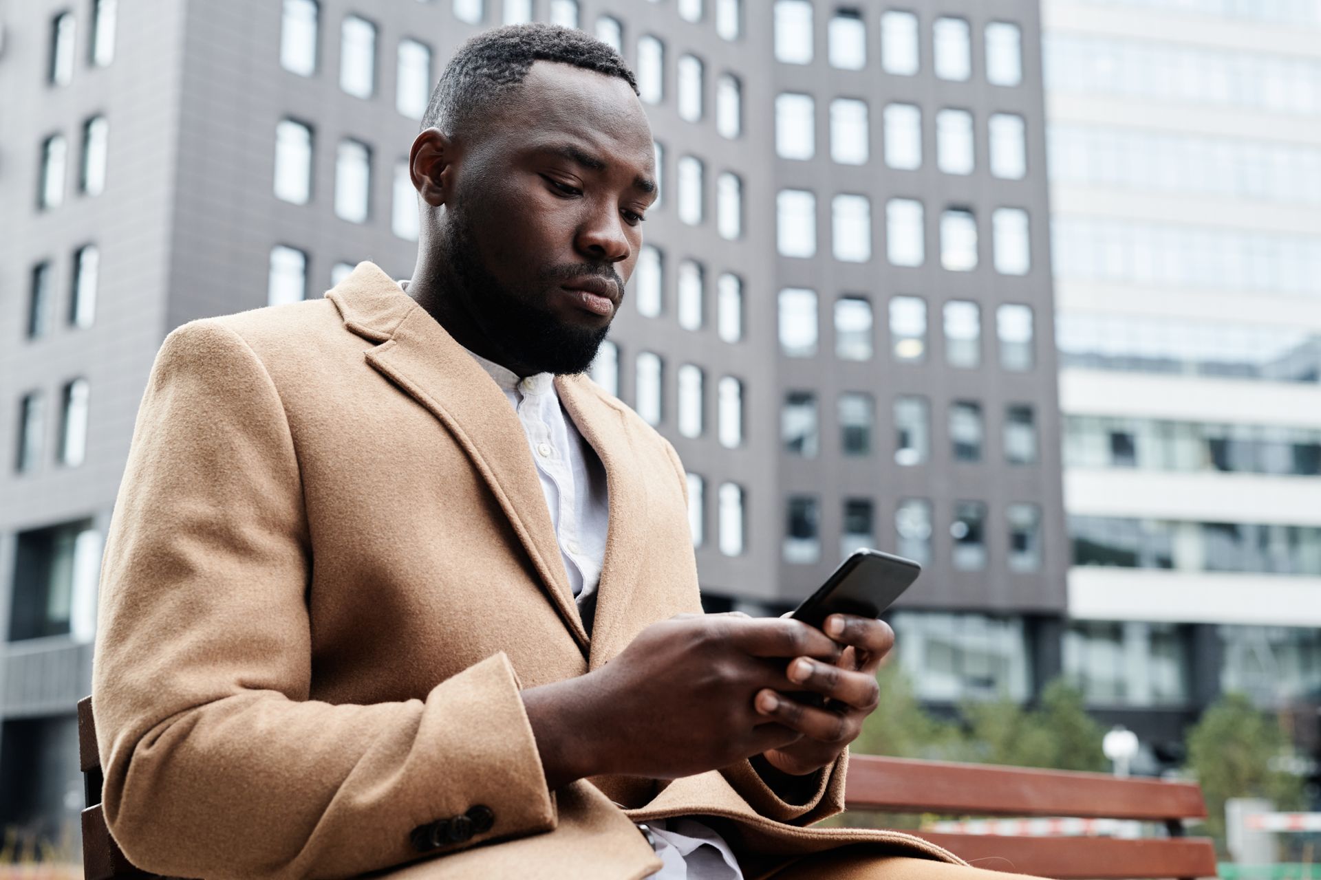 Businessman using smartphone outdoors in city environment wearing beige coat.