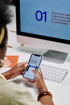 Close-up of a person using a smartphone in a tech-focused office environment, displaying content on a desktop monitor.