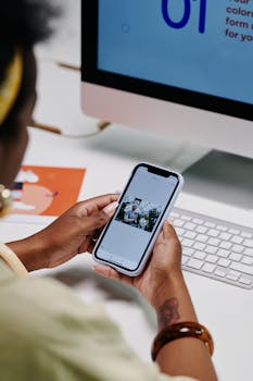 Close-up of hands holding a smartphone while browsing photos, with computer in the background.