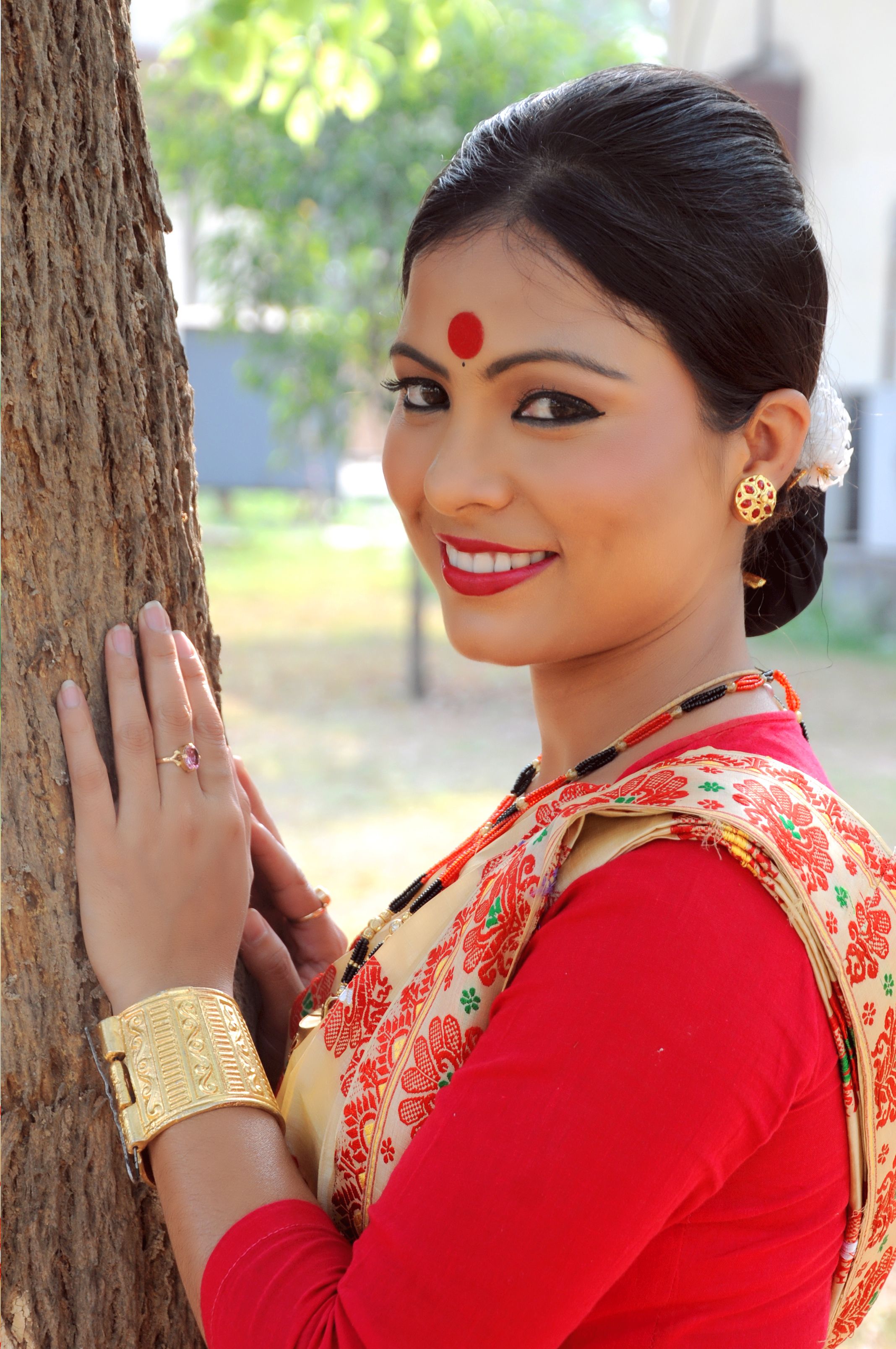 Portrait of a smiling woman in traditional South Asian attire with floral patterns, leaning against a tree outdoors.