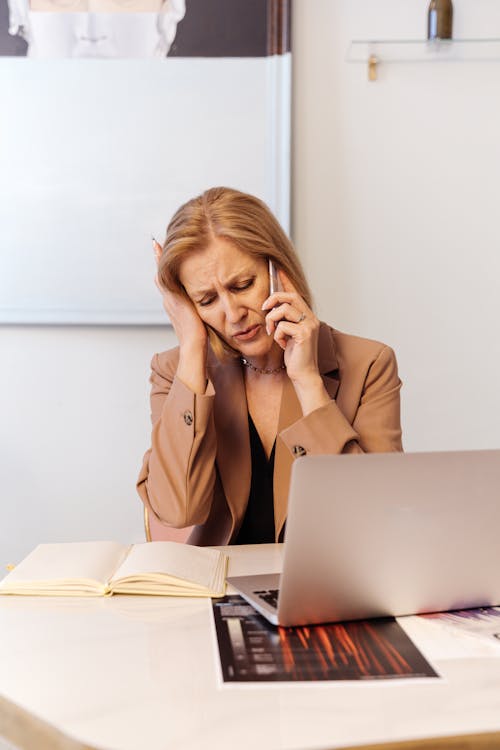 Free A Woman in Brown Blazer Having a Phone Call while Using a Laptop Stock Photo