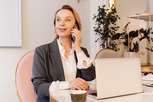 Businesswoman in blazer using smartphone while working on laptop at office desk.