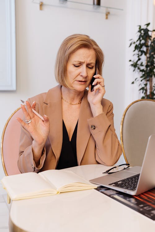 Free A Woman in Brown Blazer Having a Phone Call while Using a Laptop Stock Photo