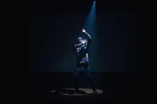 A captivating hip-hop dancer in colorful attire performs under a spotlight in a dark setting.