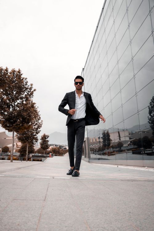 Free A stylish man in formal attire and sunglasses walking confidently by a modern glass building. Stock Photo